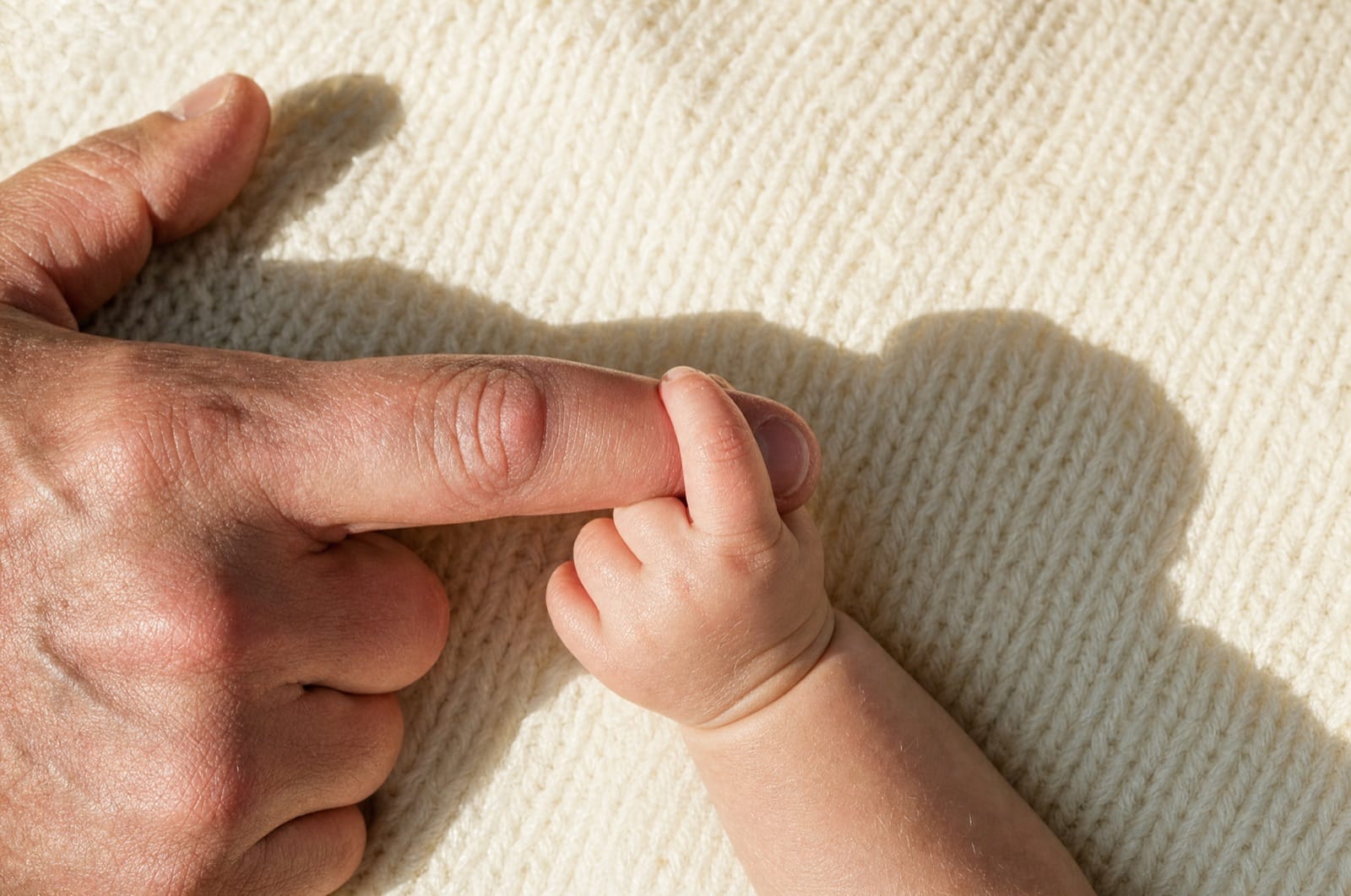 Kinderhand umgreift den Finger eines Erwachsenen auf einer hellen Wolldecke